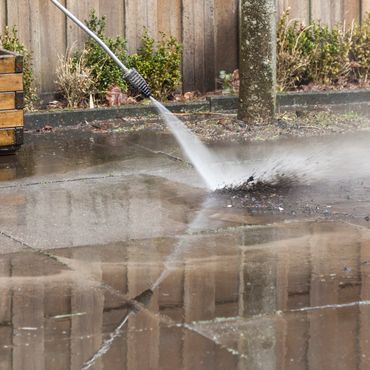 Pressure washing a dirty concrete patio near a wooden fence and plants.