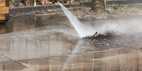 Pressure washing a dirty concrete patio near a wooden fence and plants.