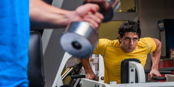 Man in yellow shirt using gym equipment while another person lifts a dumbbell.