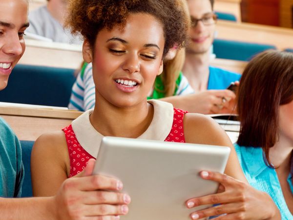 Students in a lecture hall sharing a tablet and smiling.