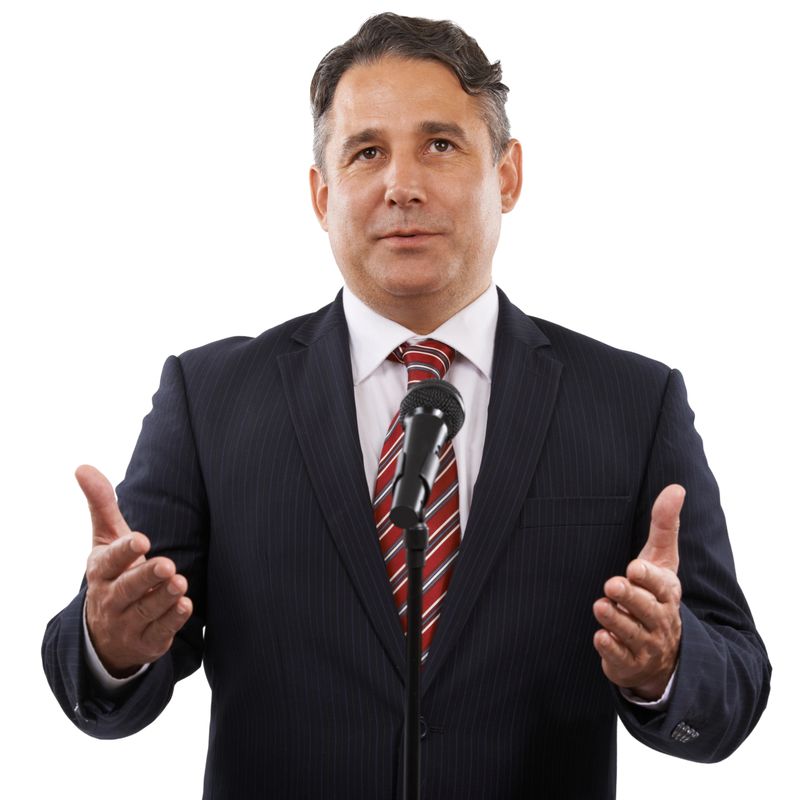 A man in a suit with his hands raised while saying a speech into a microphone