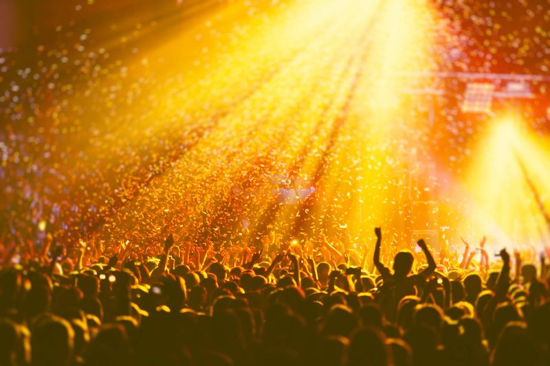 A crowded concert hall with scene stage orange and yellow lights, rock show performance, with people silhouette, colourful confetti explosion fired on dance floor air during concert festival