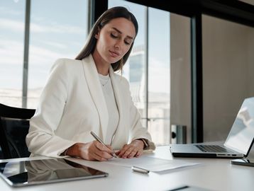 Professional woman in white blazer working at a desk with laptop and tablet.