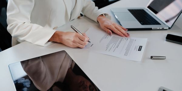 A person in white signs a contract at a desk with a laptop and tablet.