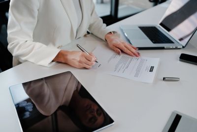 Person signing a document at a desk with a laptop and a tablet.