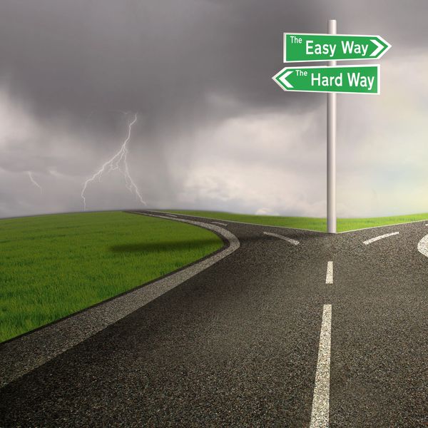 A forked road with signs pointing to 'The Easy Way' and 'The Hard Way' under stormy skies.