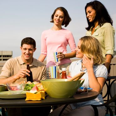 Four friends enjoying a sunny outdoor meal with drinks and food on a table.