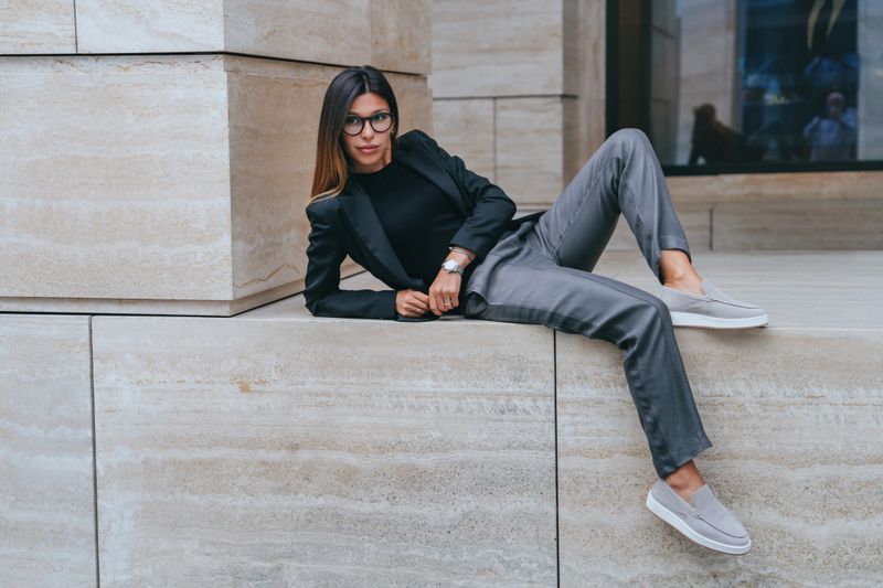 Stylish young brunette woman in black jacket and grey pants posing outside laying looks away with confident face expression. Mockup, fashion, successful women. Model in city.