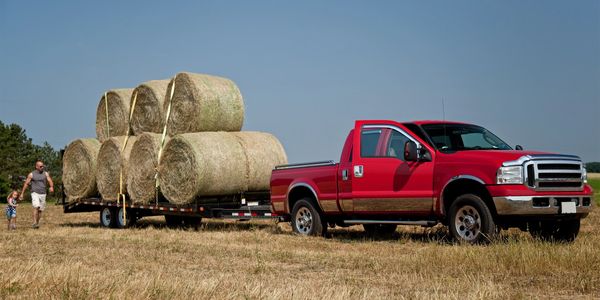 A red pickup truck towing a trailer loaded with large hay bales in a field.