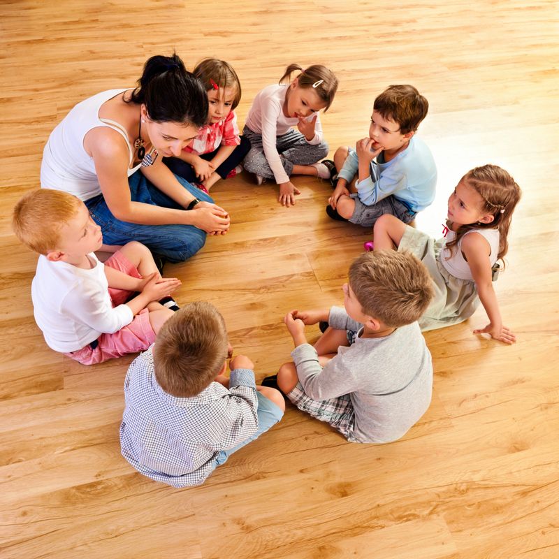 Nursery school children sitting in circle on the floor in a playroom and listening to their teacher. High angle view.