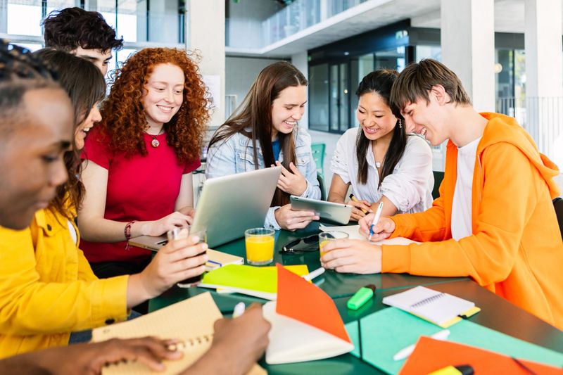 Young group of high school students studying together at cafeteria campus table. Diverse young people with books and laptops doing group study. Education and teamwork concept.