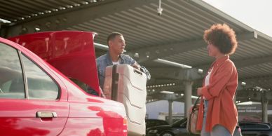 Man lifting suitcase from car trunk talking to woman.