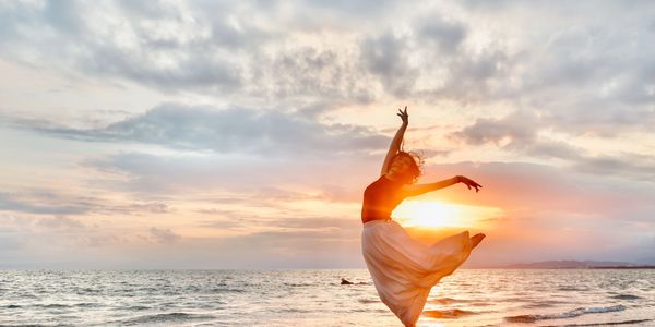 A woman dances joyfully on the beach at sunset.