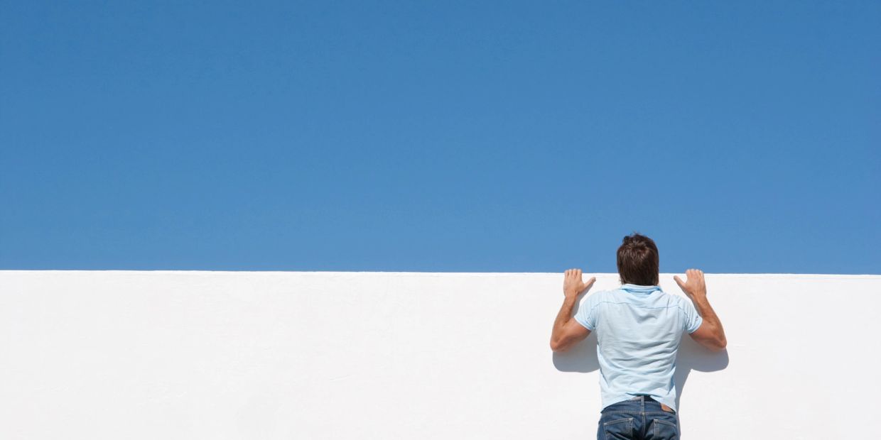 Man peeking over a white wall under a clear blue sky.