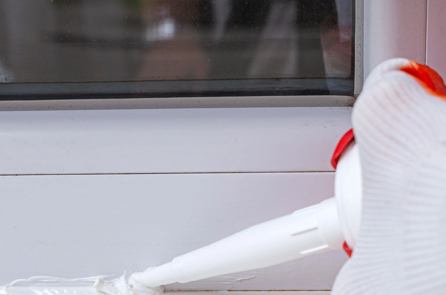 Gloved hand applying white sealant to window frame gap.