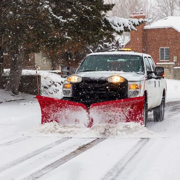 A truck with a red snowplow clearing snow on a residential street.