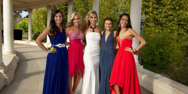 Five young women in elegant evening gowns posing outdoors at a formal event.