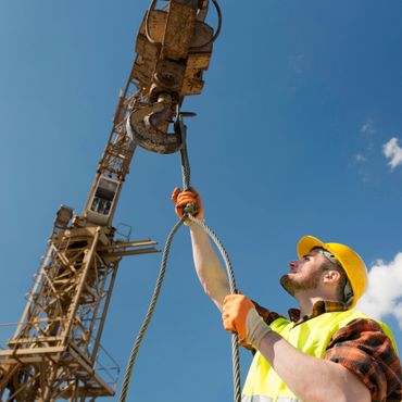 Construction worker securing a cable to a crane hook under a clear blue sky.
