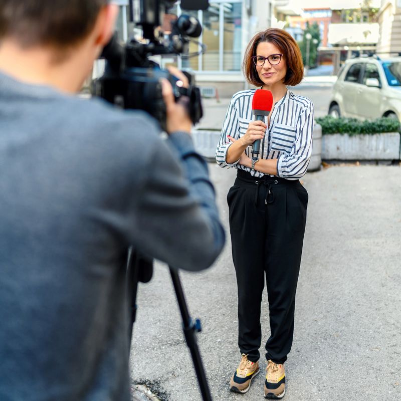 Female television reporter holding a microphone, she is standing outdoors in front of a building. Male camera operator is filming her.
