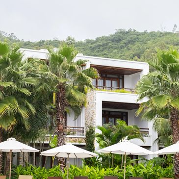 Resort with pool, sun loungers, umbrellas, and lush greenery against a mountain backdrop.