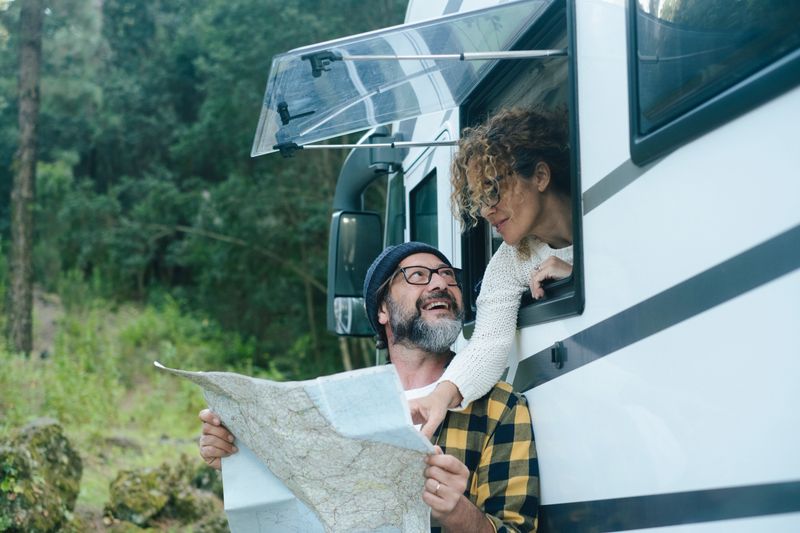Adult couple use map guide to choose next travel destination enjoying freedom and vacation outside a modern camper van. Nomadic lifestyle people. Man and woman traveler fulltime in nature green park