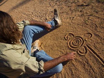 A man with long hair draws a spiral pattern in the dirt while sitting.