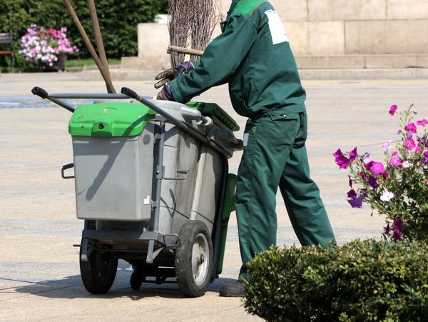 Worker in green uniform pushing a large waste bin outdoors.