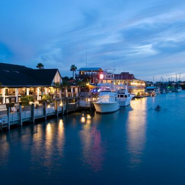 Evening view of a marina with boats docked and waterfront buildings lit up.
