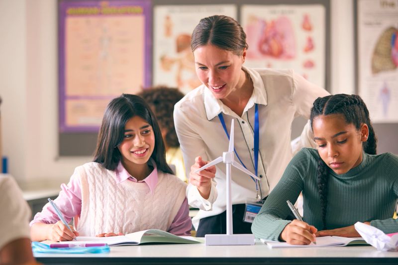 Female Teacher Helping Secondary Or High School Students Studying Wind Turbines In Science Class