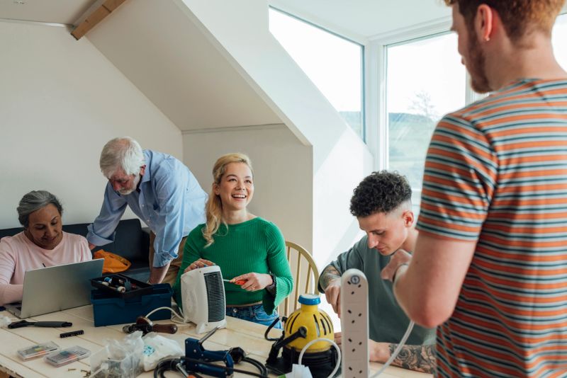 A group of people taking part in a repair shop in a community room in Horsley, Northumberland. They are sitting round a wooden table and learning to fix different technology equipment while a senior man helps them all and passes on his knowledge. The main focus is a young woman smiling and looking at her friend.
