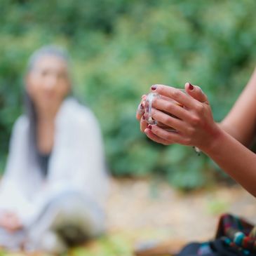 A woman holding a clear cup, with others sitting blurred in the background outdoors.
