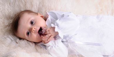 A baby in a white outfit lying on a fluffy surface, holding a pacifier.