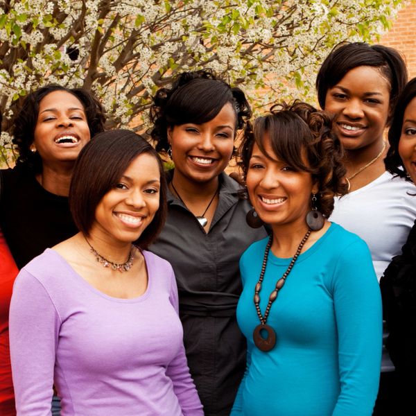 A group of six smiling women standing outdoors near flowering trees.