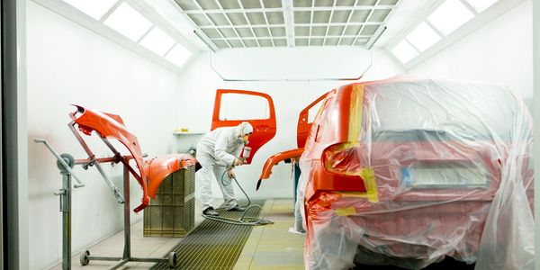 Worker spray-painting a red car and parts in a paint booth.