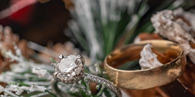 Close-up of a diamond engagement ring and a gold wedding band on snowy pine needles.