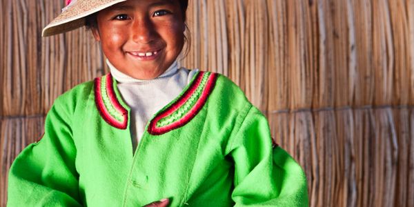 Smiling child in traditional green attire and hat against a rustic backdrop.