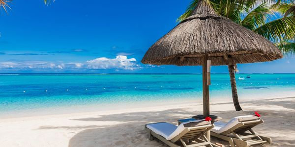 Two lounge chairs under a straw umbrella on a tropical beach with clear blue water.