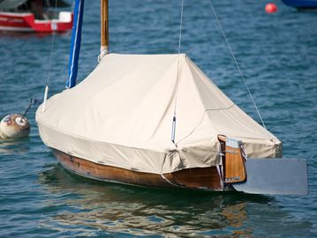 A small wooden sailboat covered with a beige tarp floating on calm water.