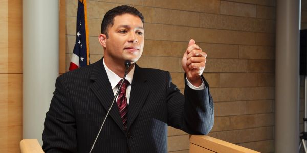 Man in suit speaking confidently at a podium with an American flag behind him.