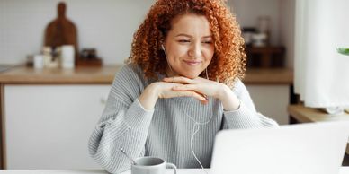 A woman with curly red hair engages with her laptop, smiling gently while wearing earphones.