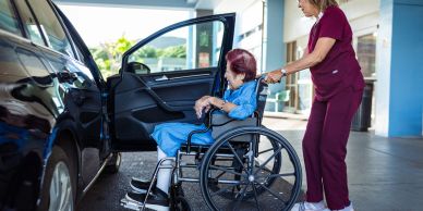 Caregiver pushing elderly woman in wheelchair towards car.
