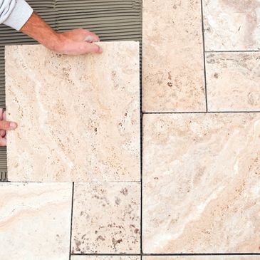 Hands installing beige stone tiles on a floor.
