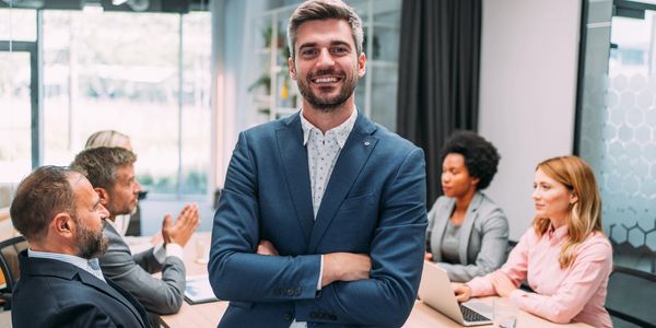 Confident businessman standing with arms crossed in a meeting room with team members.