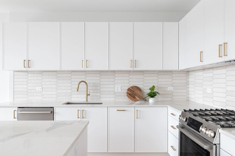 A beautiful kitchen detail with white cabinets, a gold faucet, white marble countertops, and a brown picket ceramic tile backsplash.