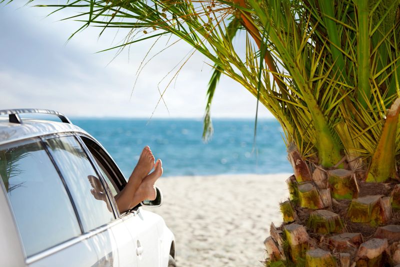 Woman hanging legs out of car window by the palm tree on tropical sandy beach.