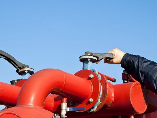Hand turning a valve on a large red industrial pipe against a clear blue sky.