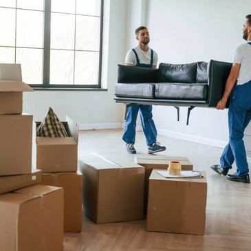 Two movers carrying a black couch in a room with packed boxes.