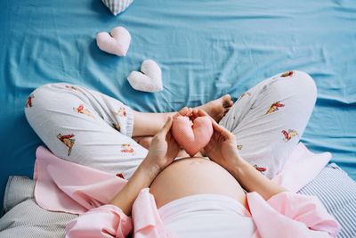 Pregnant woman holding a heart-shaped pillow on her belly, sitting on a bed.