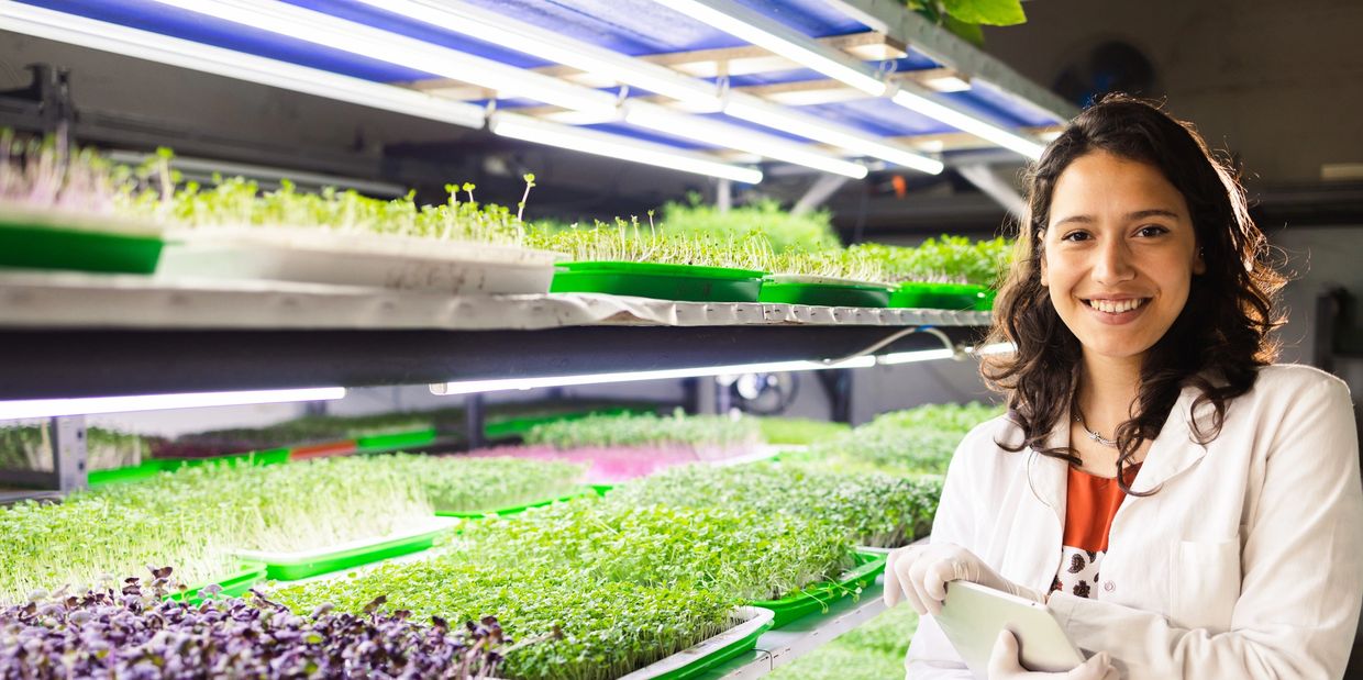 Scientist in lab coat smiles next to trays of microgreens under grow lights.