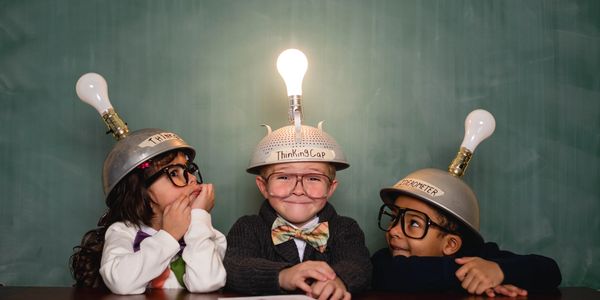 Three children wearing creative thinking caps with light bulbs, sitting at a table.